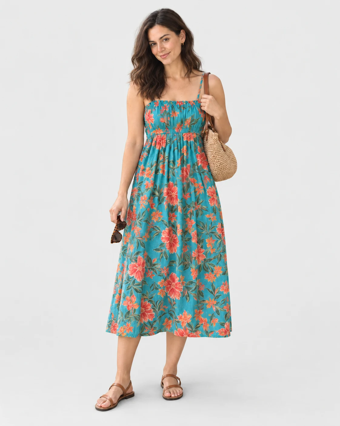The same woman now wears the turquoise floral dress, tan sandals and gold hoops against a light grey studio background, carrying the raffia bag and sunglasses.